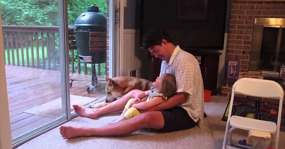 A man sits on the floor holding a baby, with a dog lying nearby, near a sliding glass door.