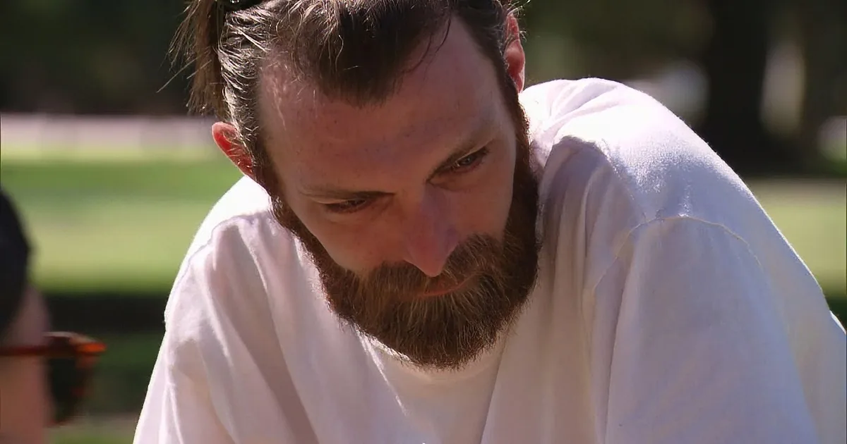 A man with a beard and white shirt is looking down, possibly in thought or concentration.