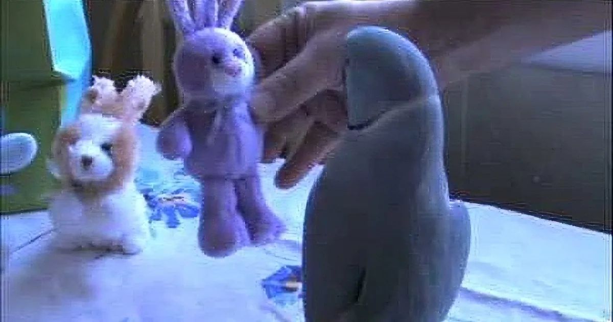 A hand holds a purple stuffed bunny, surrounded by other plush toys on a table.