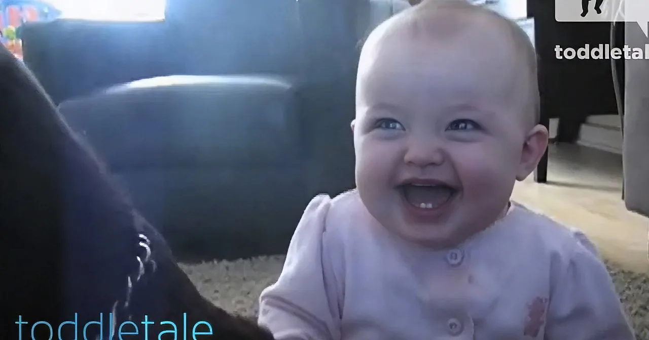 A baby in a pink onesie is smiling widely, with a dark object partially obscuring the lower part of the image.