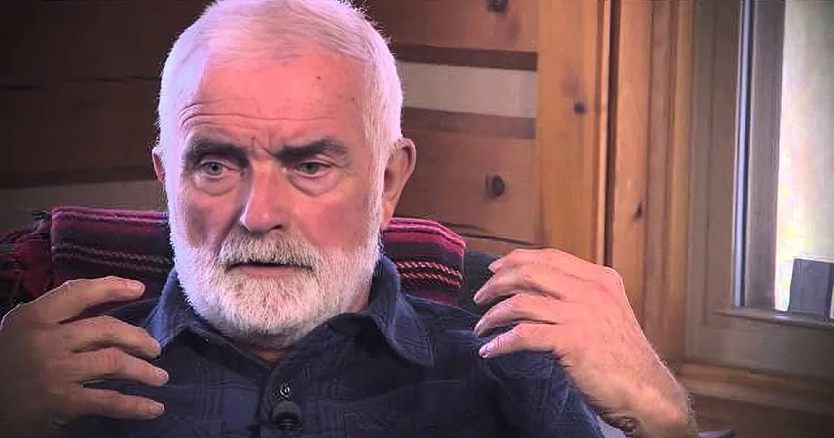 An older man with white hair and beard sits in a chair, gesturing with his hands while speaking.