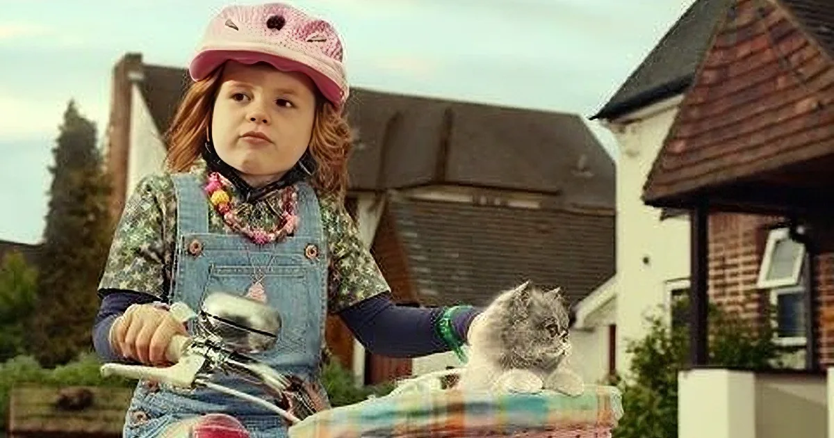 A young girl in a pink helmet and denim overalls rides a bicycle with a basket, surrounded by a suburban neighborhood.