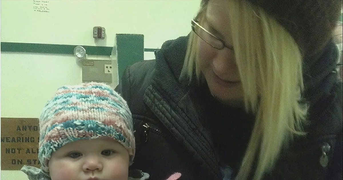 A woman with blonde hair and glasses holds a baby wearing a striped hat in a room with a sign in the background.