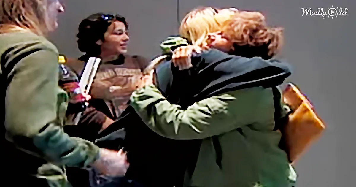 A person in a green jacket is hugging another person in a black outfit, with a third person holding a book in the background.