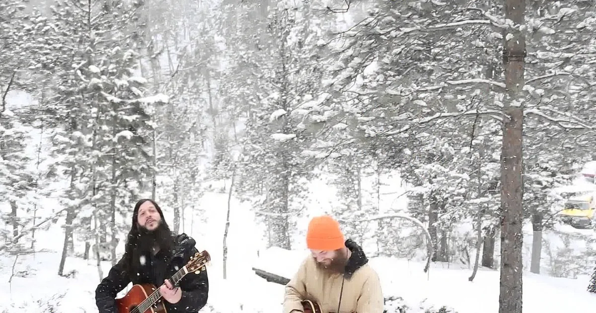 Two individuals play music in a snowy forest setting, surrounded by snow-covered trees.