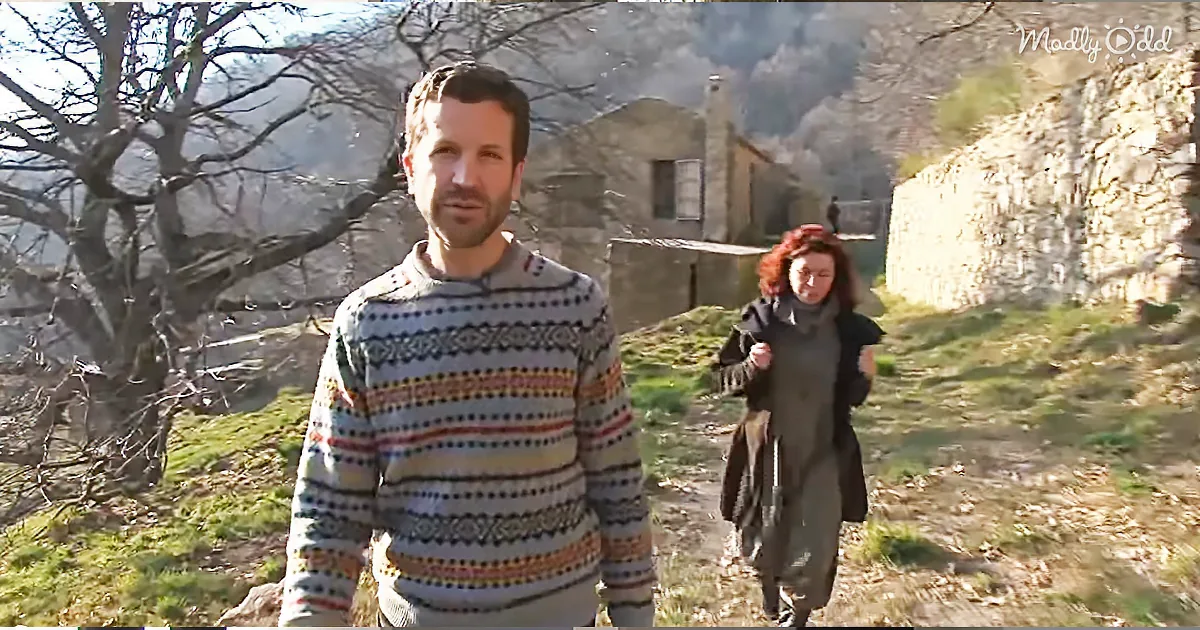 A man in a patterned sweater stands in a rustic setting with a woman walking behind him.