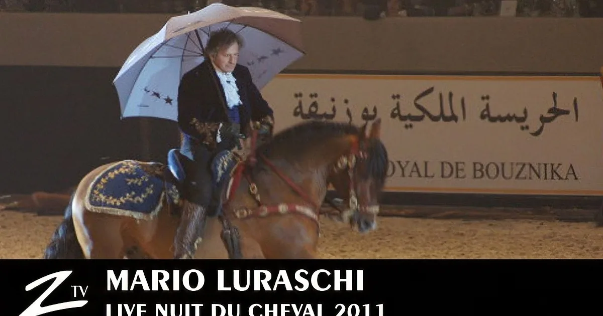 A man in formal attire rides a horse under an umbrella in an arena.