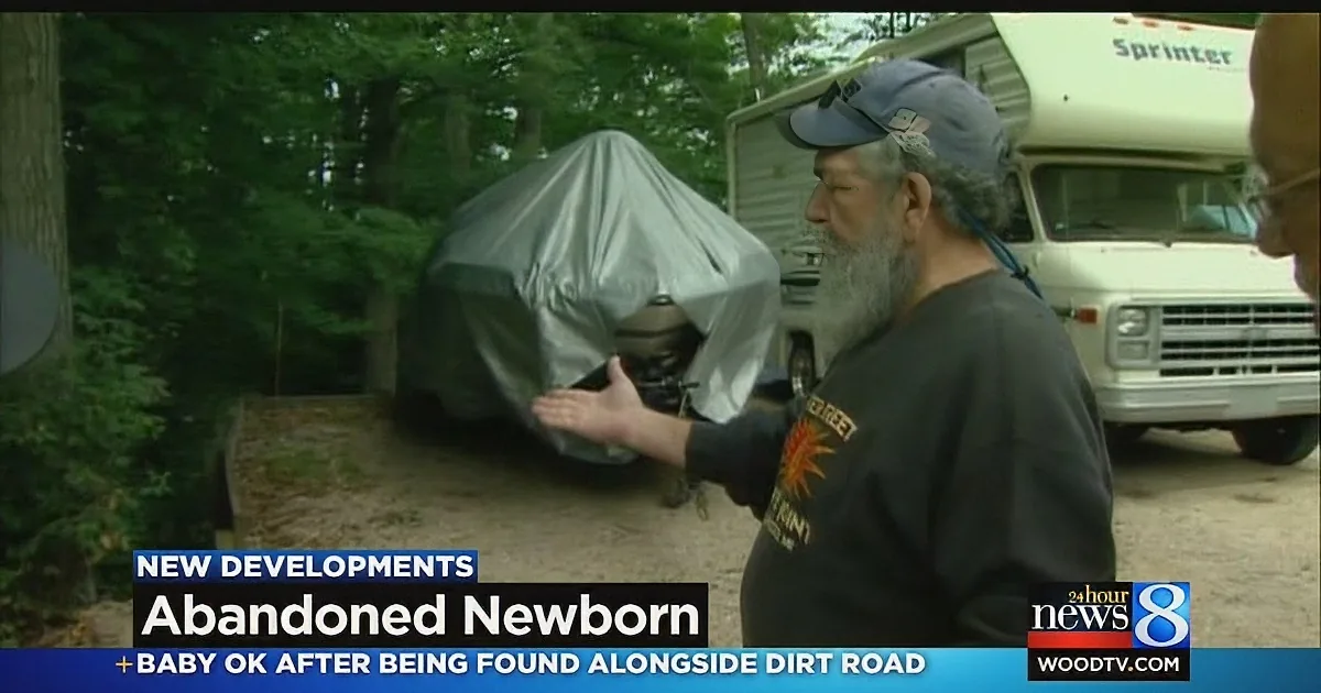 A man with a beard and cap gestures near a covered object, with an RV and trees in the background.