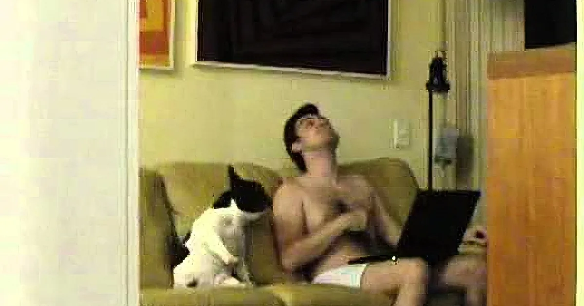 A shirtless man sits on a couch, leaning back, with a black and white cat sitting on his lap.