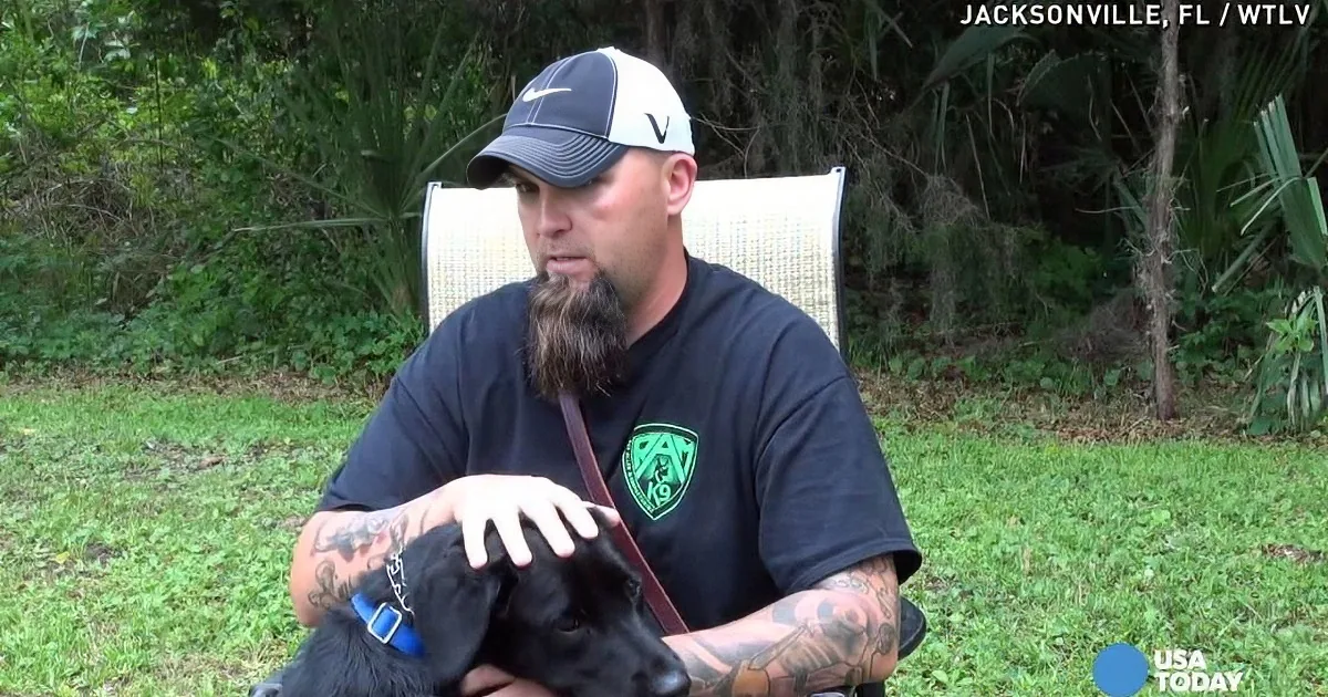 A man with tattoos and a beard sits outdoors, holding a black dog with a blue collar.