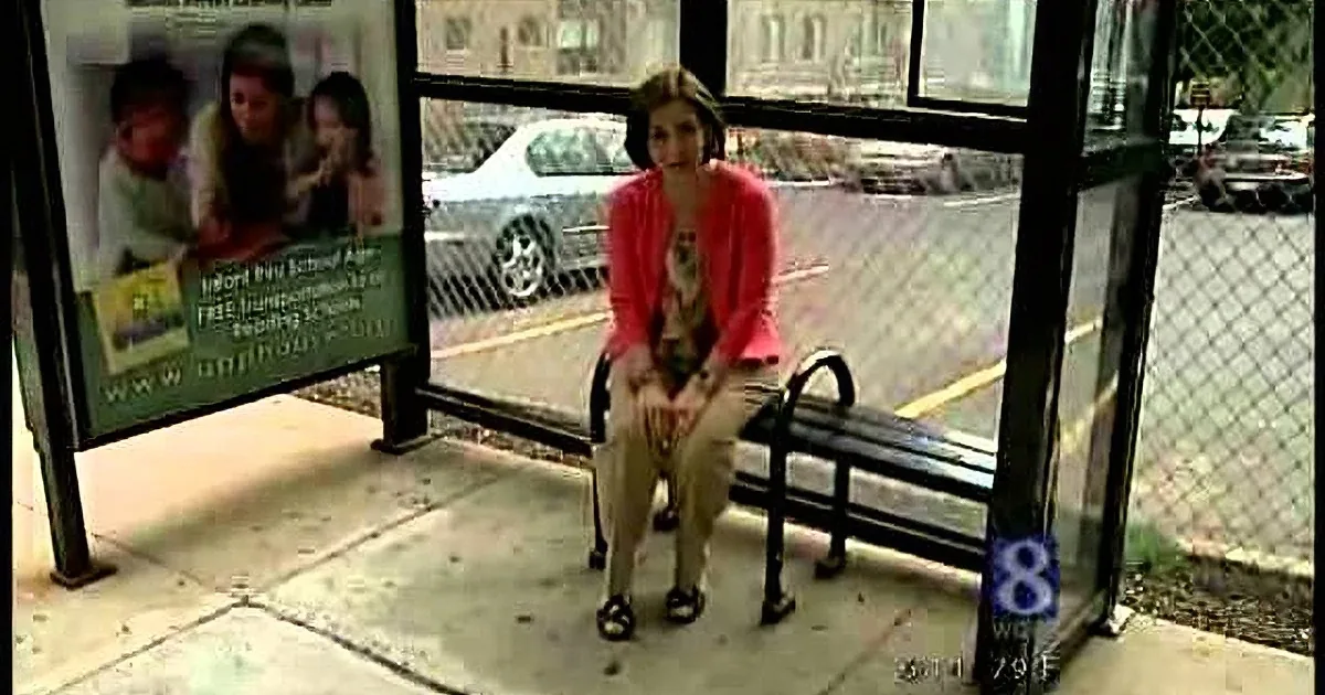 A woman in a red cardigan sits on a bench at a bus stop, surrounded by advertisements and a chain-link fence.
