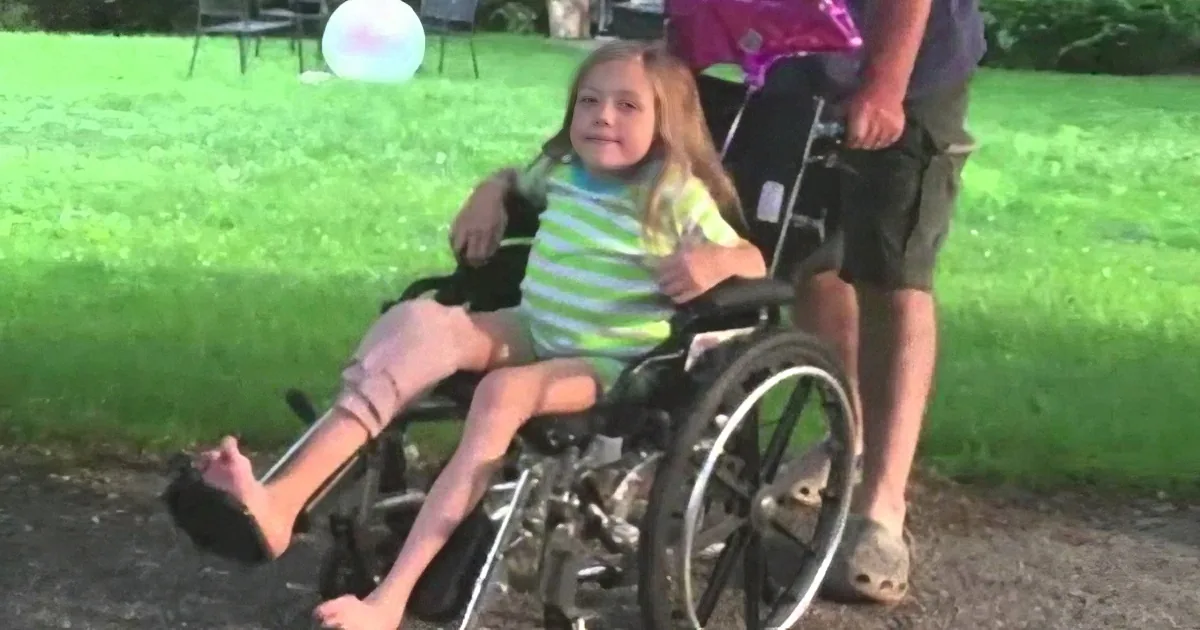 A young girl in a wheelchair smiles as a man pushes her on a grassy field.