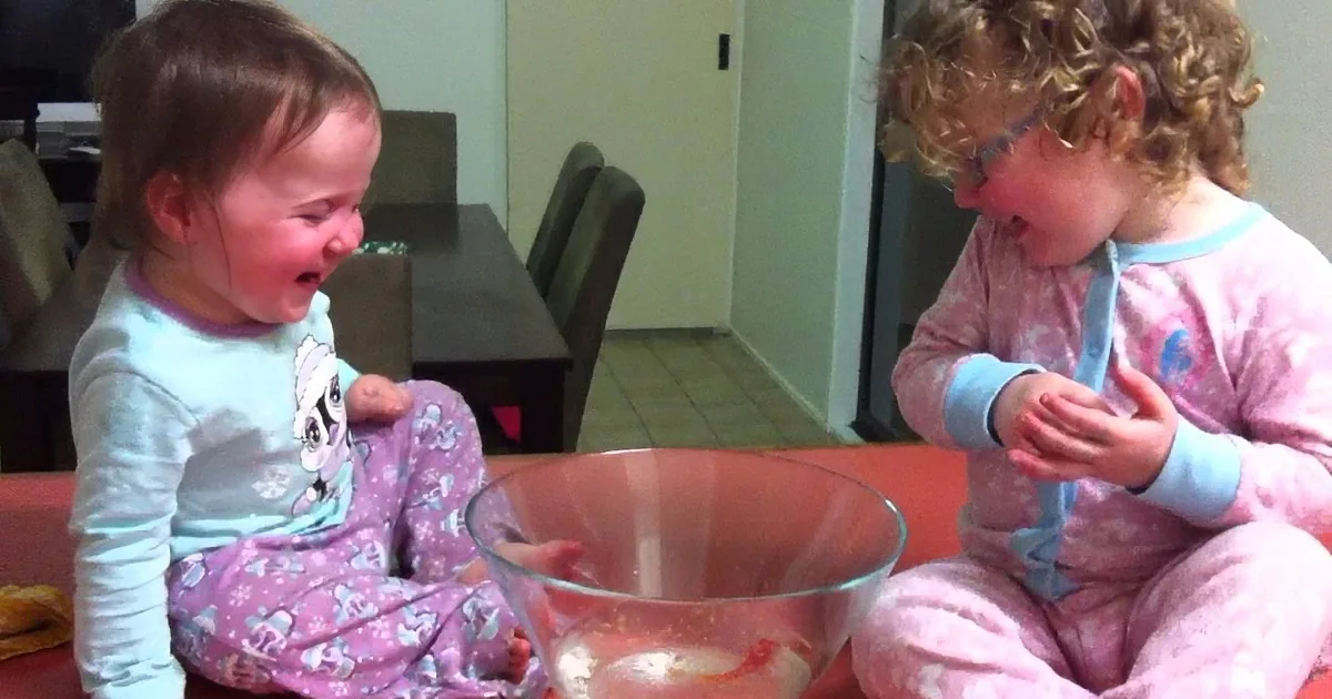 Two children in pajamas sit at a table, one crying, the other eating, with a glass bowl in front of them.
