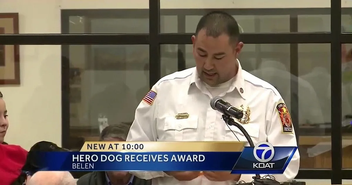 A man in a white uniform speaks at a podium, with a banner below stating "Hero Dog Receives Award."