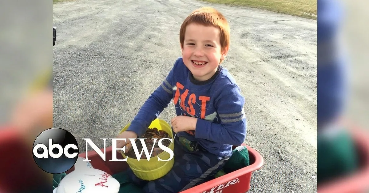 A young boy smiles in a red wagon, holding a yellow bucket, with a gravel road and grass in the background.