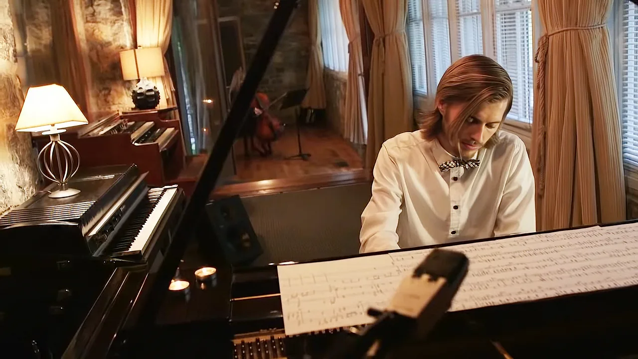A man in a white shirt and bow tie plays a grand piano in a warmly lit room with music sheets and a cello in the background.
