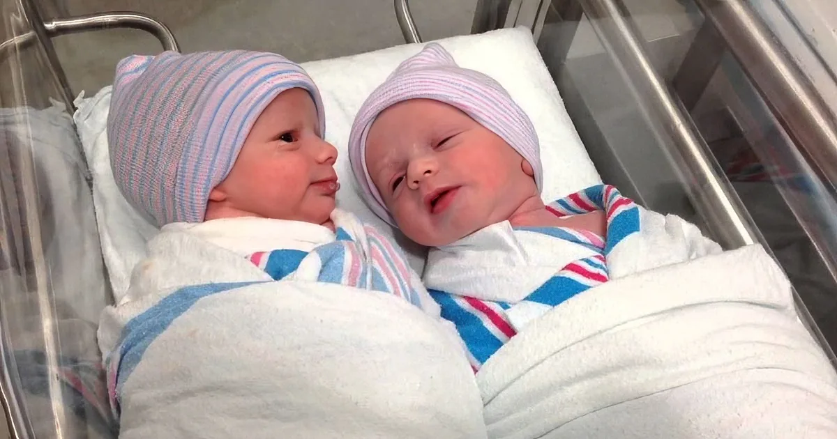 Two newborn babies wrapped in blankets and wearing striped hats lie side by side in a hospital bassinet.