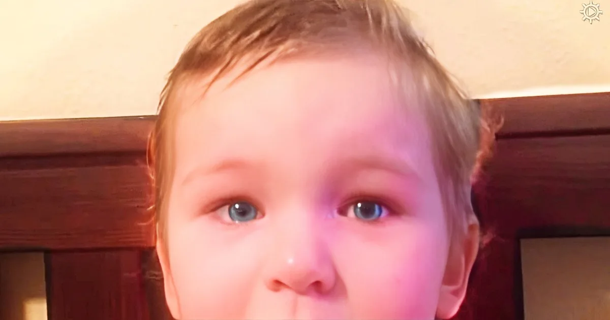 A young child with light skin, blue eyes, and short hair, wearing a red garment, is looking directly at the camera.