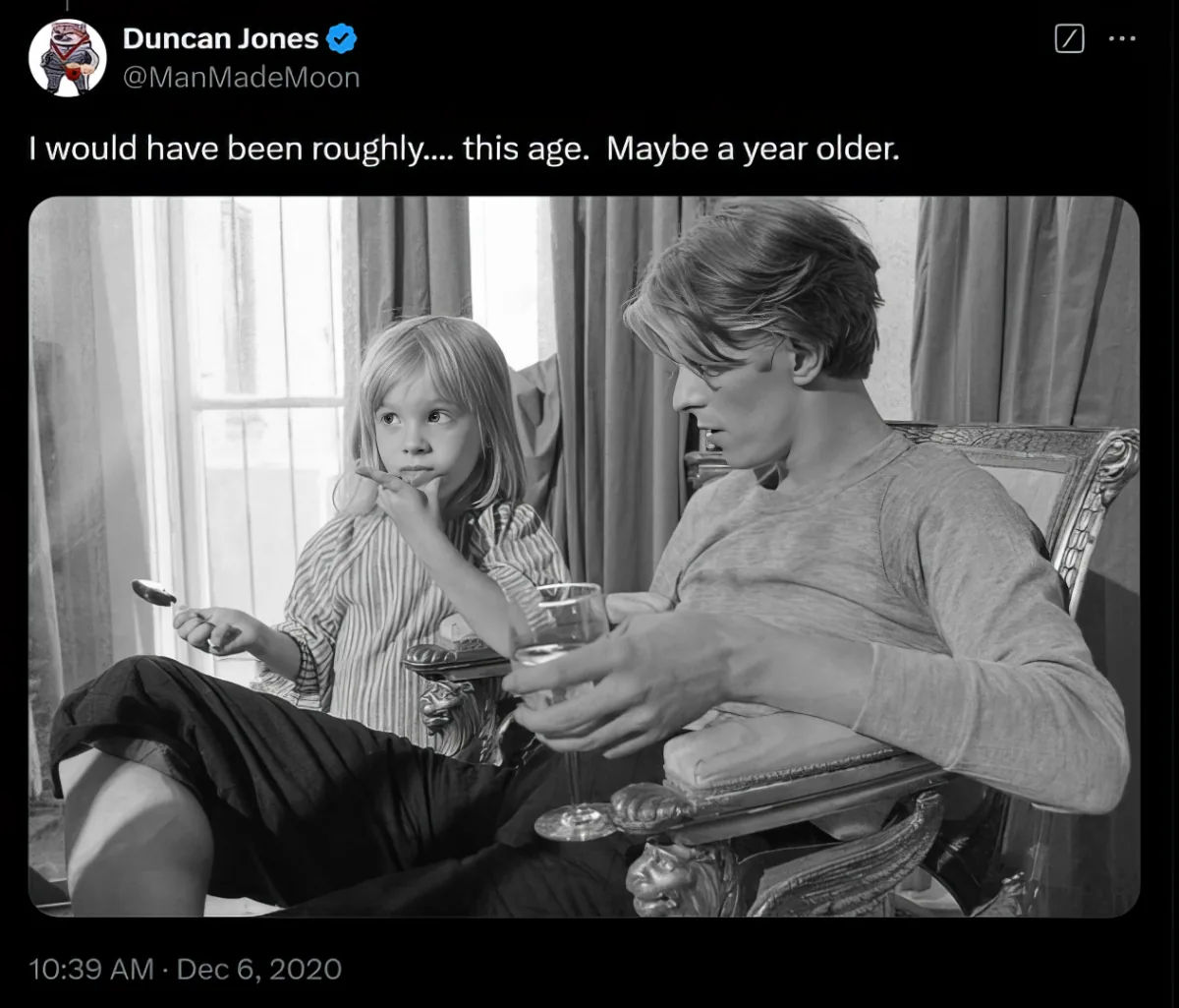 Text: A black and white photo tweeted by Duncan Jones showing a young child and David Bowie seated together, the child eating and Bowie holding a glass, illustrating Jones's age when his father filmed with Bing Crosby.
