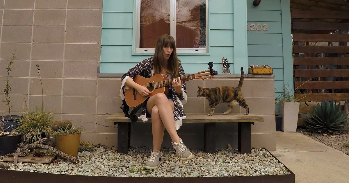 A woman plays guitar on a bench as a cat jumps nearby, with a house and plants in the background.