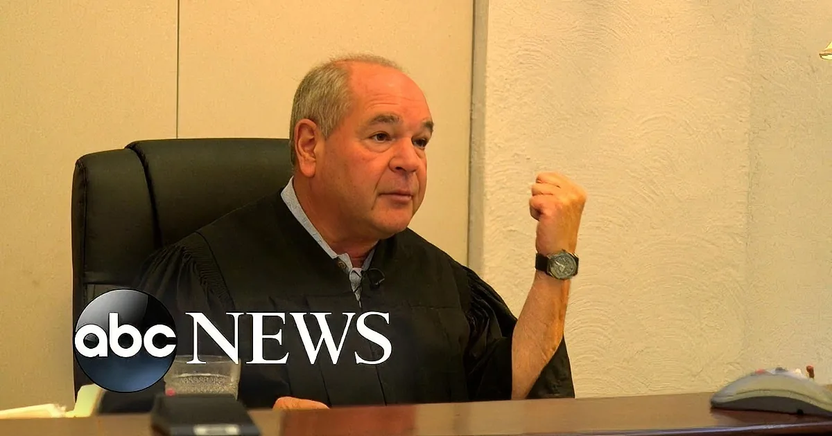 A man in a judge's robe sits at a desk, gesturing with his right hand.