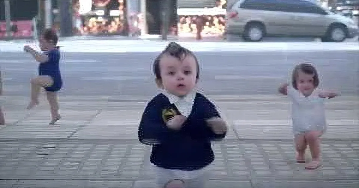 A baby in a Batman shirt stands on a sidewalk, surrounded by other children playing.