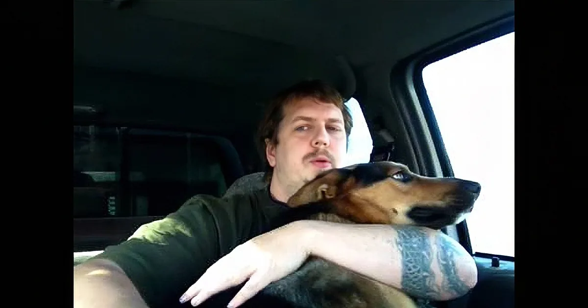A man sits in a car, petting a dog that leans on the window.