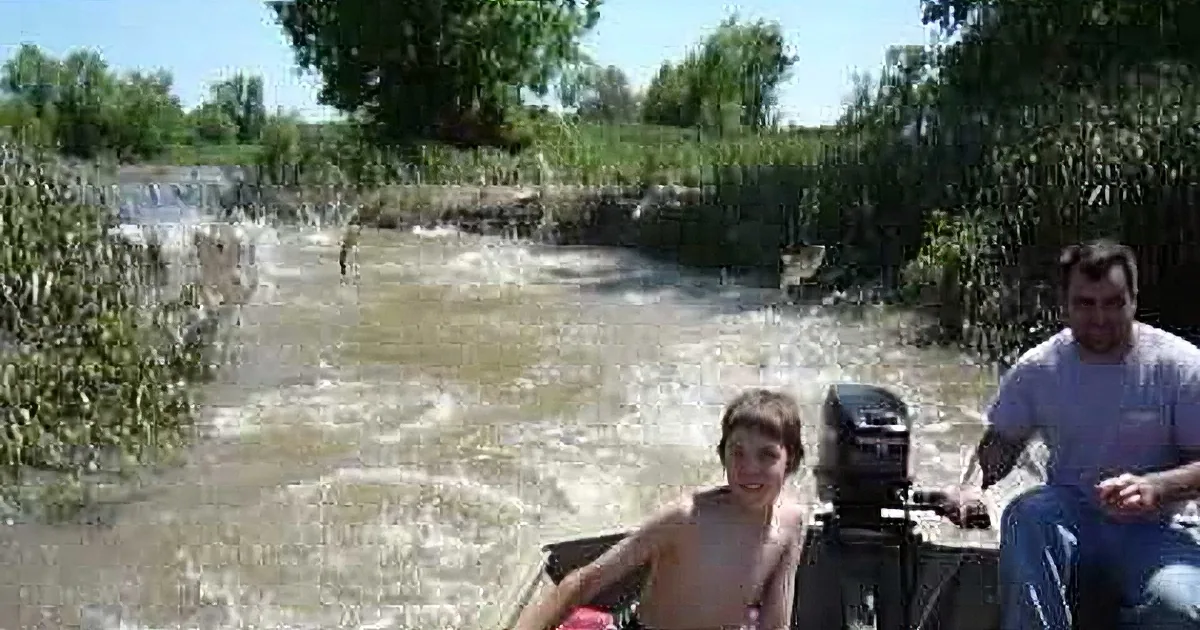 A man and child ride a boat through a fast-moving river surrounded by greenery.