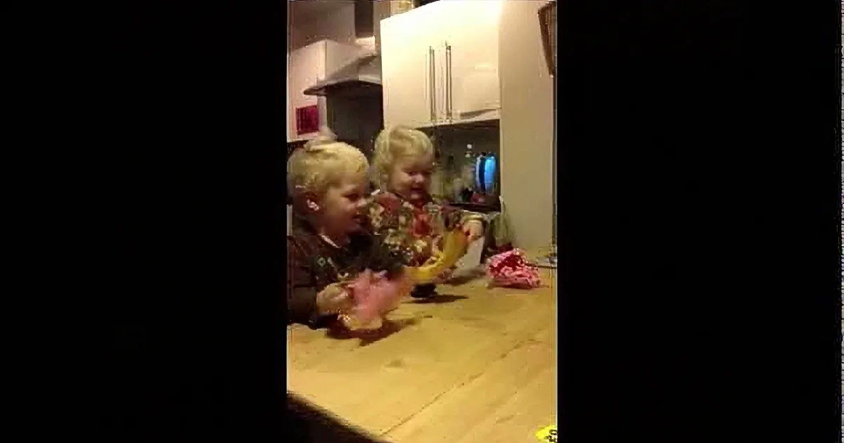 Two children are sitting at a kitchen table, one holding a pink object, the other looking at it.