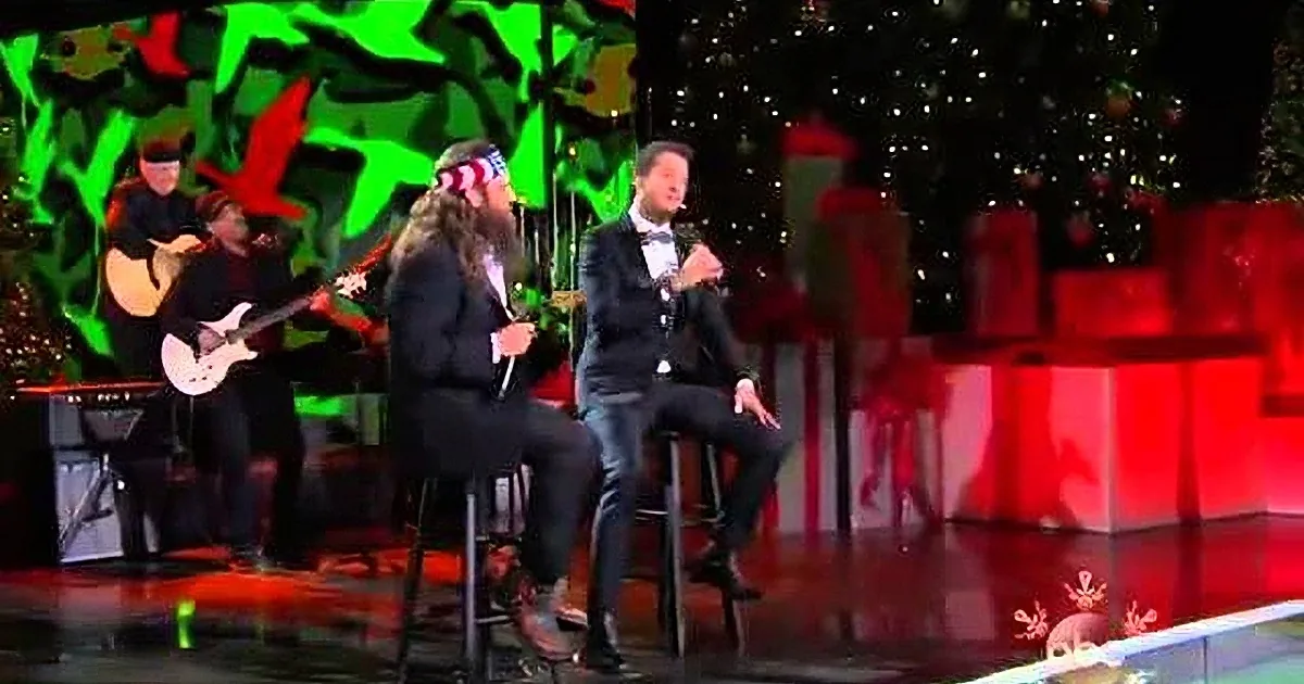 A man in a suit sings on stage with a guitarist, surrounded by festive decorations and a Christmas tree.