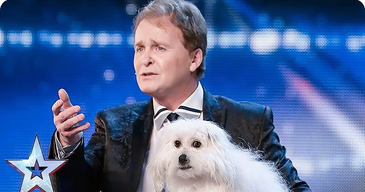 A man in a suit gestures while holding a fluffy white dog on a stage with a blue background.