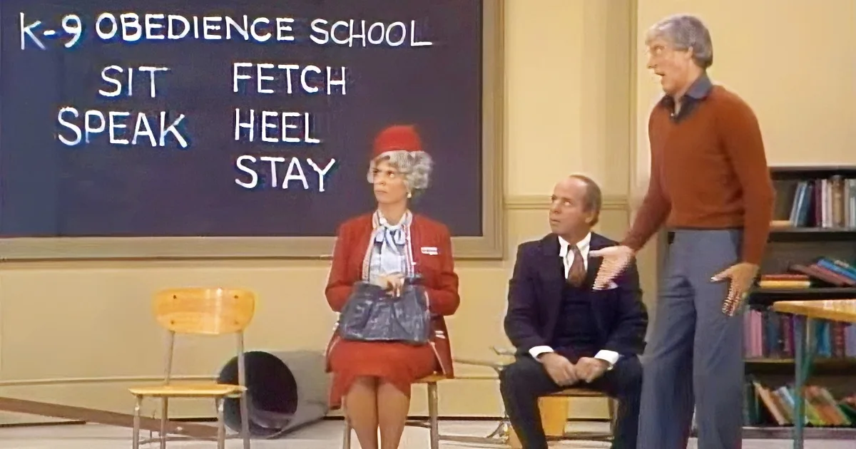 A woman in a red uniform sits on a chair, with a man in a suit and another standing nearby in a classroom setting.