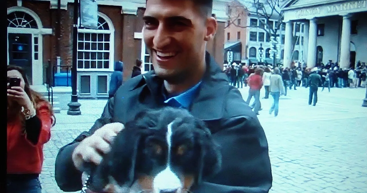A man smiles while holding a black and white dog in a bustling outdoor setting with people and buildings in the background.