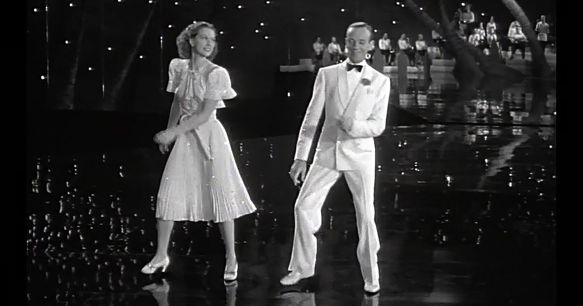 A man in a white suit and bow tie dances with a woman in a white dress on a reflective stage.