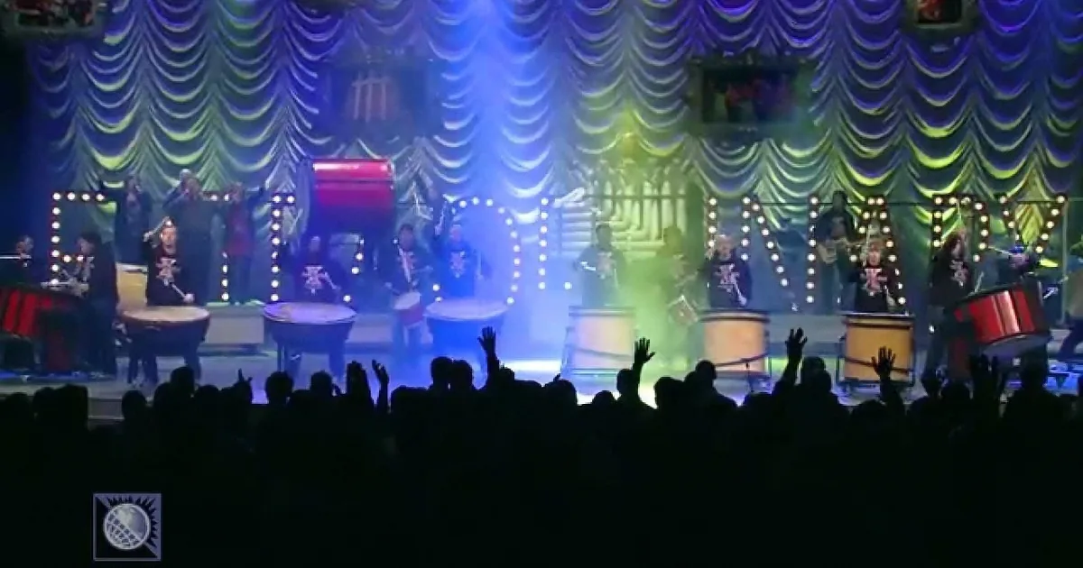 A band performs on a brightly lit stage with a crowd raising their hands in front.