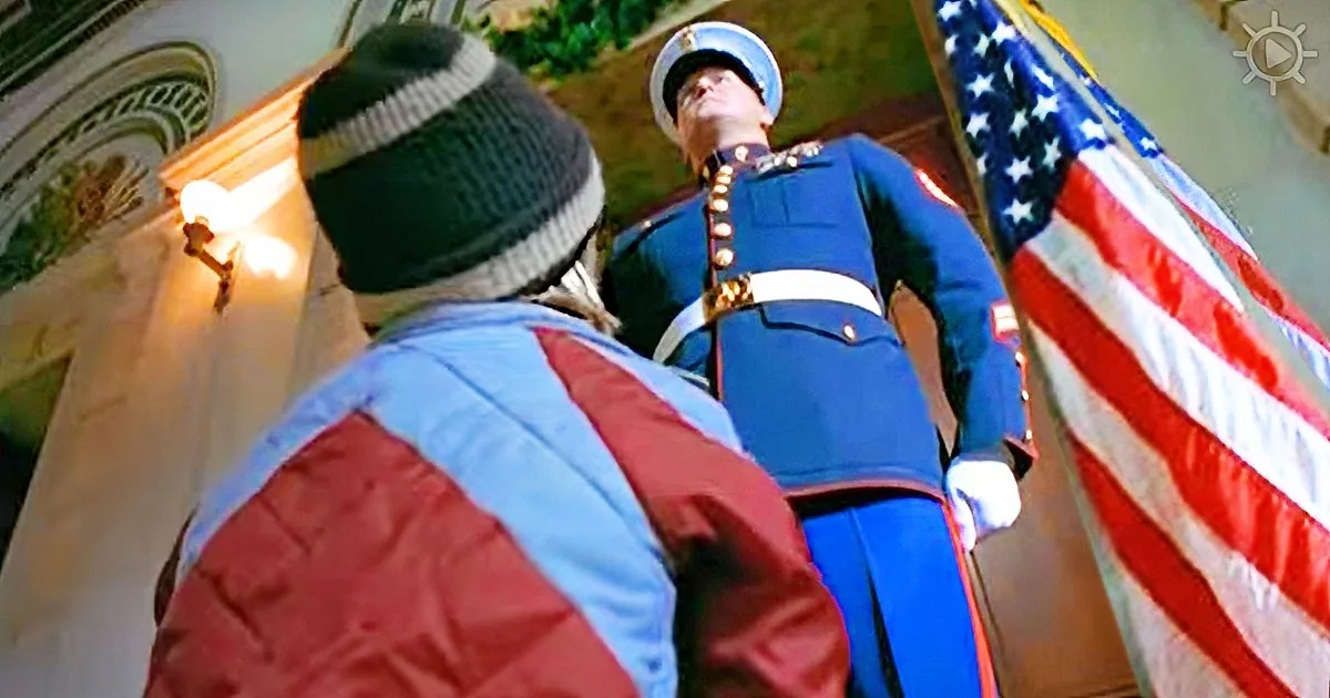 A person in a winter hat and jacket stands below a uniformed guard near an American flag.