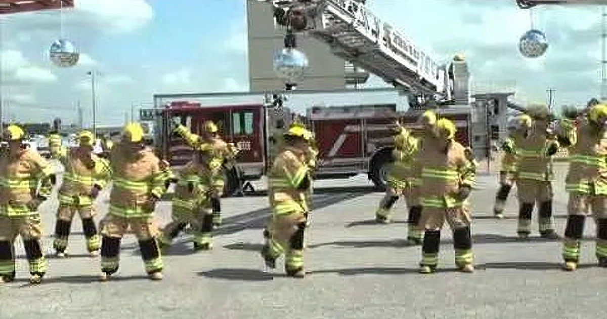 Firefighters in yellow gear perform a synchronized dance in front of a fire truck.