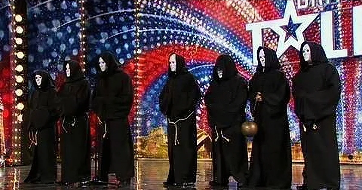 A group of seven individuals in black robes and hooded masks stand on a stage with a colorful backdrop.