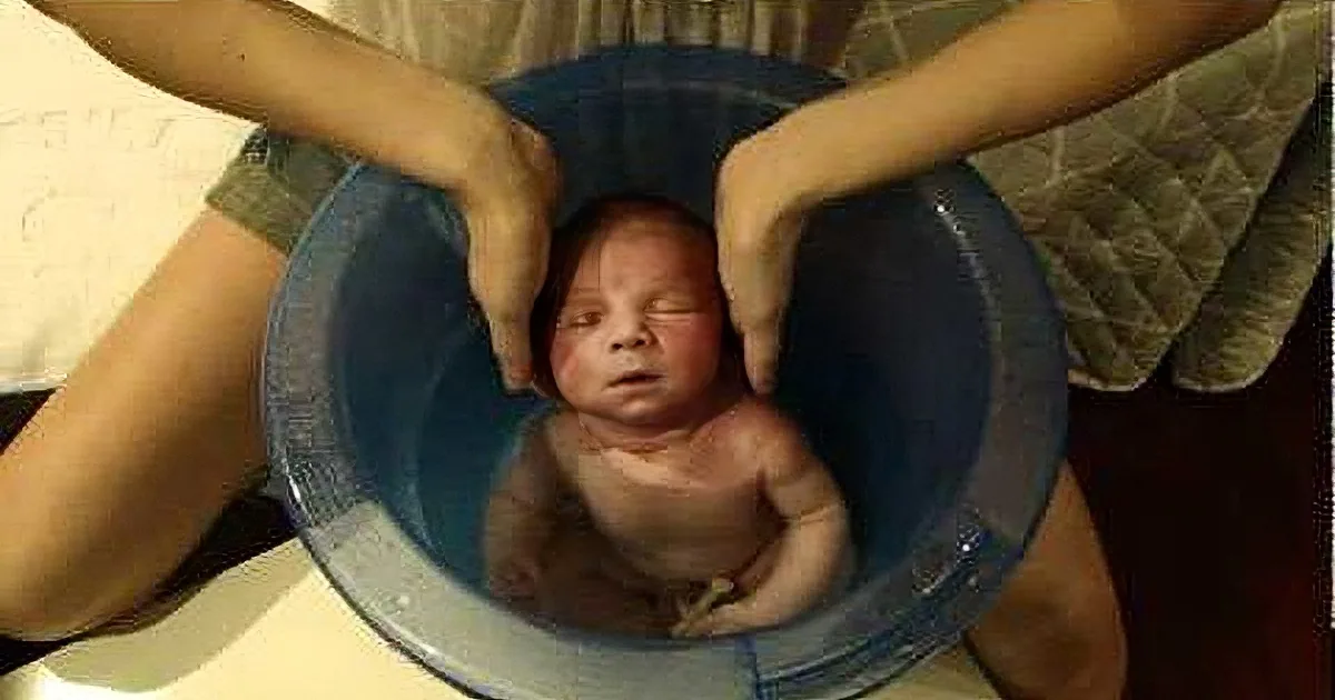 A newborn baby is being held in a blue bucket, with hands supporting the baby's head and shoulders.
