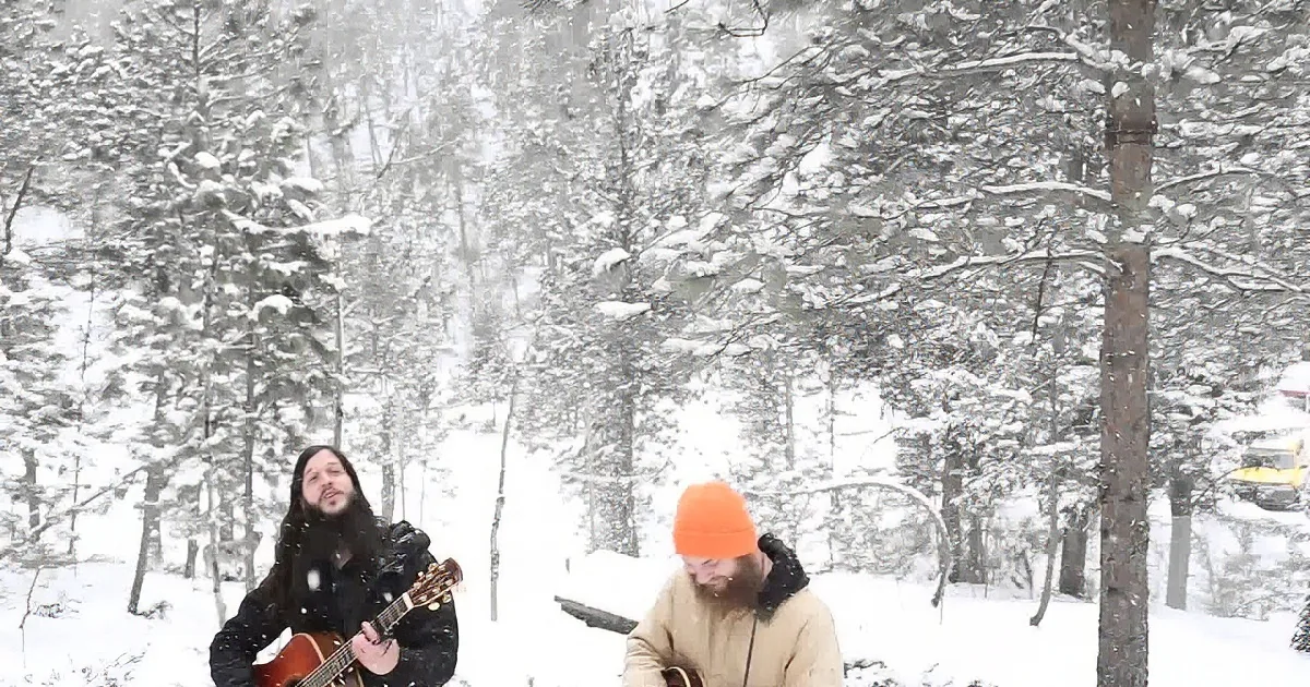 Two individuals play music in a snowy forest, one with a guitar and the other with a banjo.