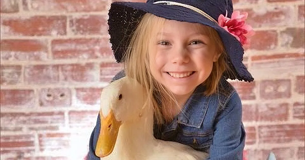 A smiling girl in a denim jacket and hat holds a duck against a brick wall backdrop.