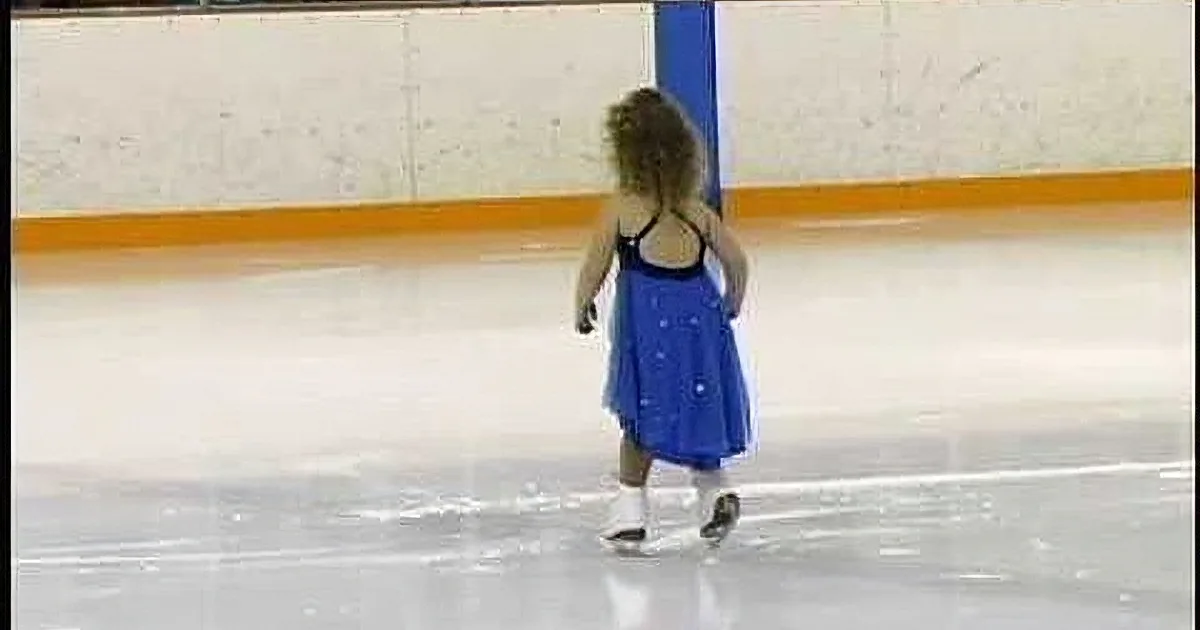 A young girl in a blue dress skates on an ice rink, her hair flowing.
