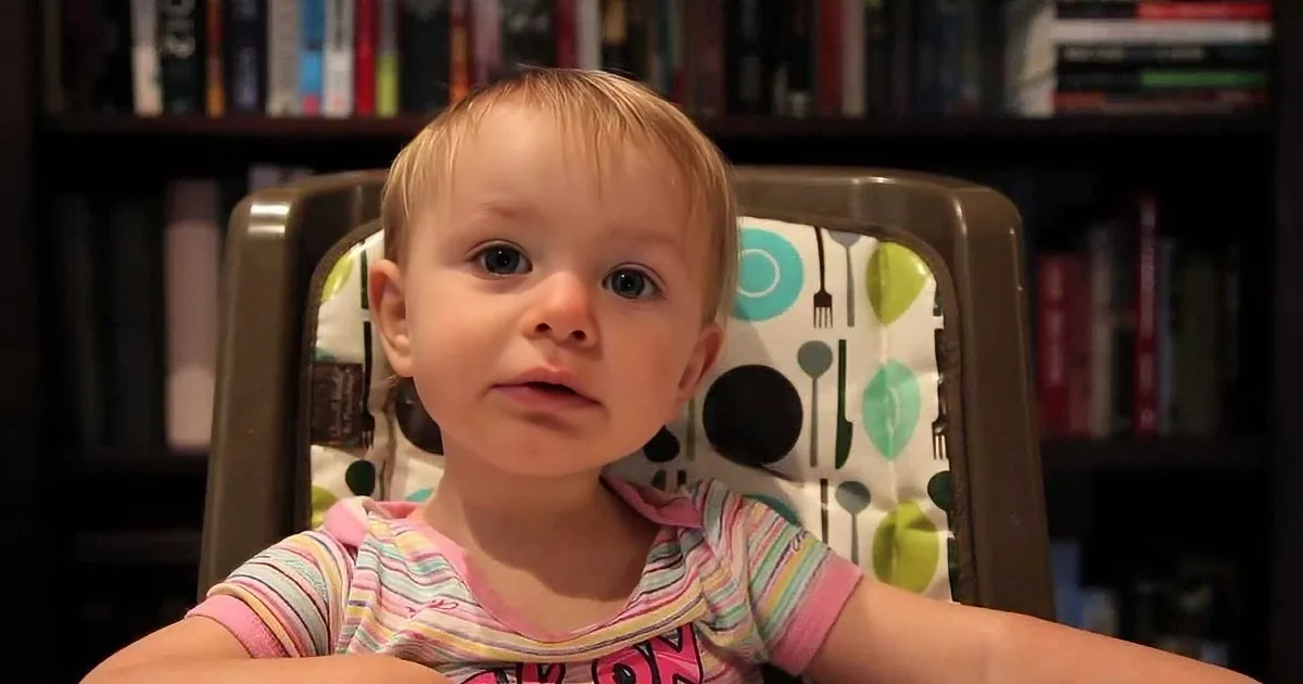 A baby in a striped shirt sits in a high chair with a colorful patterned backrest, looking forward.