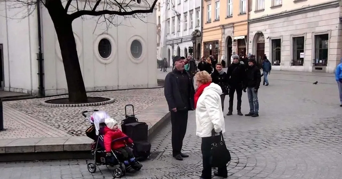 A woman in a red coat interacts with a child in a stroller on a cobblestone square with people and buildings in the...