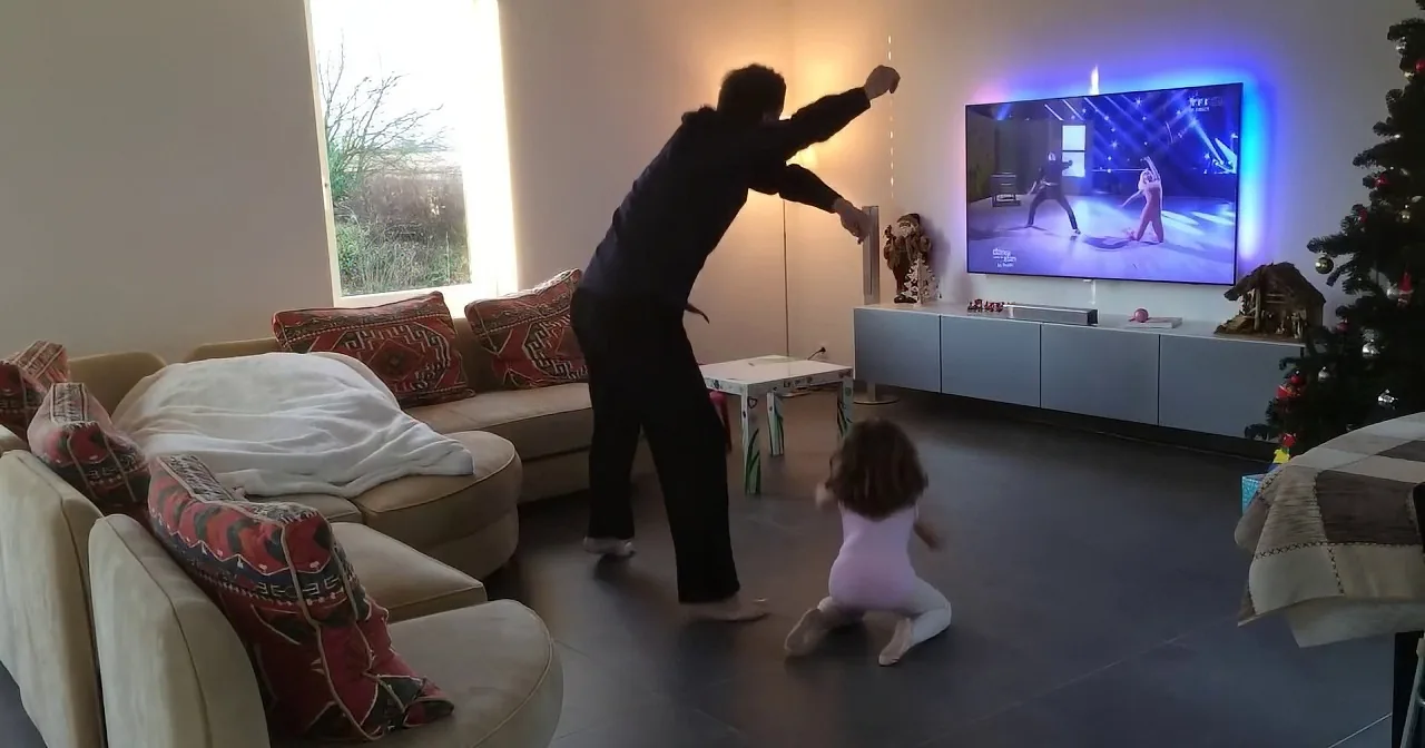 A person is mid-air, mimicking a martial arts move in a living room with a Christmas tree and a TV showing a similar scene.