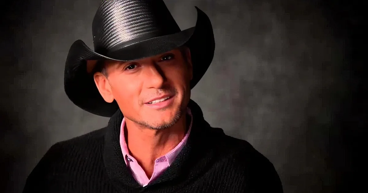 A man wearing a black cowboy hat and a black sweater over a pink shirt is smiling.