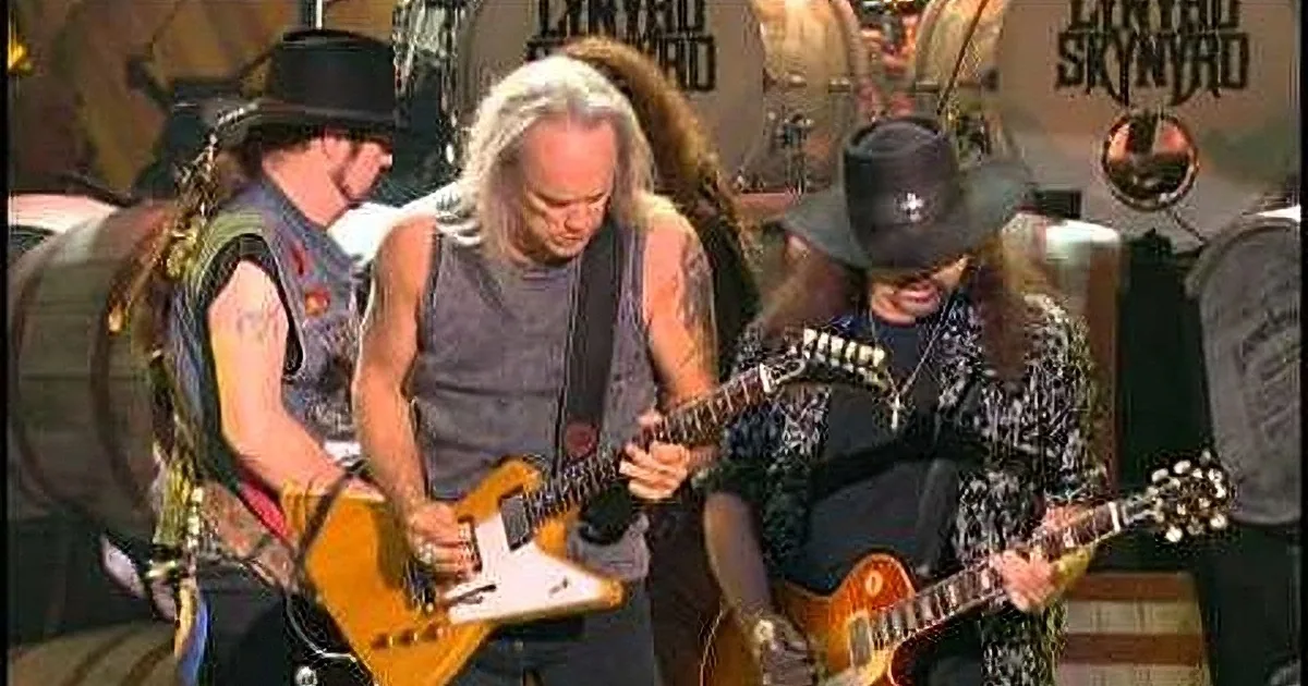 A guitarist in a black tank top and cowboy hat plays a white electric guitar on stage with other musicians.