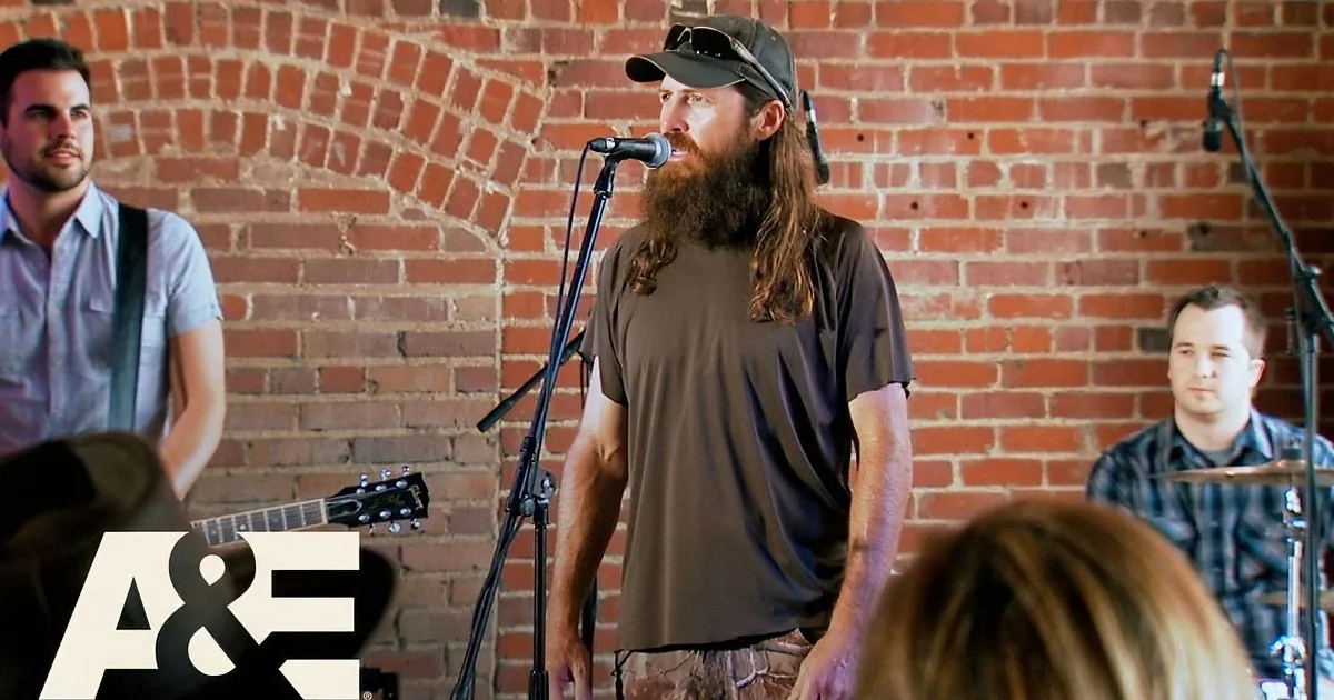 A man with a beard and cap sings into a microphone in front of a brick wall, accompanied by musicians.