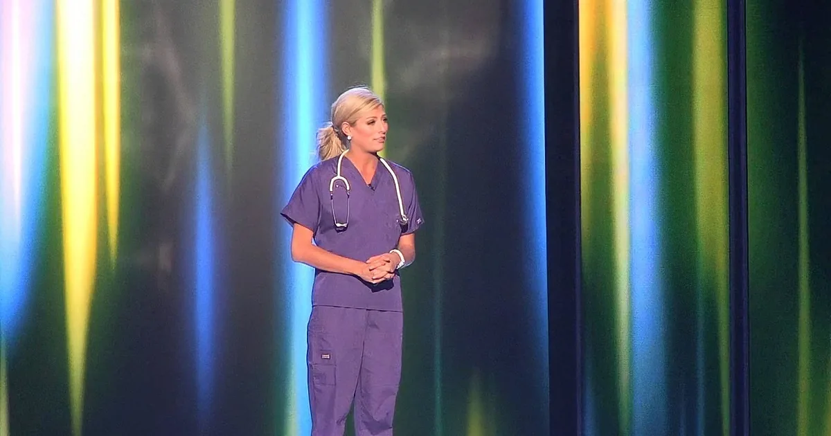 A woman in purple scrubs stands on stage with a stethoscope, against a colorful, striped backdrop.