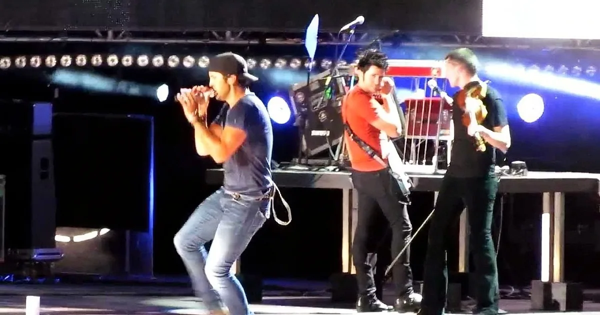 A man in a blue shirt and cap is singing into a microphone on stage, surrounded by other musicians.